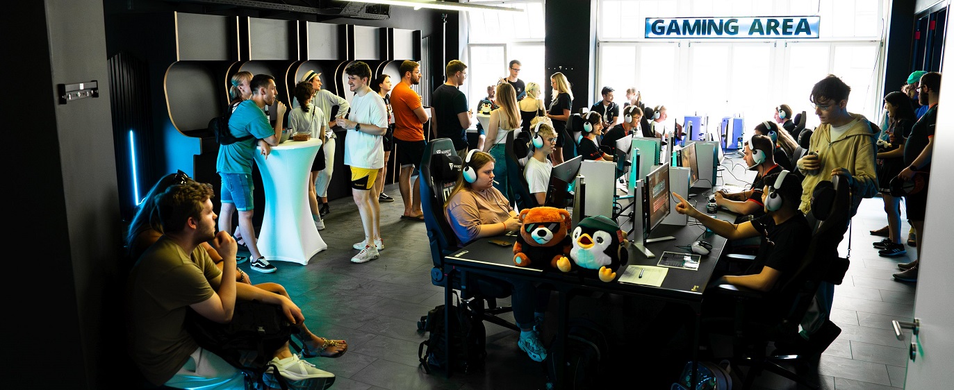 Viele Menschen befinden sich in einem Raum mit der Aufschrift Gaming Area, in dem sie sich entweder unterhalten oder an Schreibtischen mit Computern sitzen
