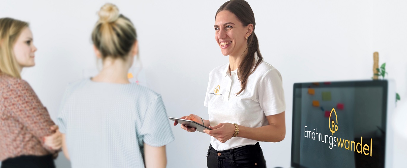 Friedericke Kirsten, Gründerin von Ernährungswandel, berät mit einem Tablet in der Hand zwei Frauen, daneben sichtbar ein großer Monitor mit dem Logo von Ernährungswandel