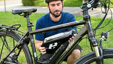 Symbolbild des Unternehmens Liofit: Ein Radfahrer hockt hinter seinem Fahrrad und hält einen E-Bike-Akku in den Händen
