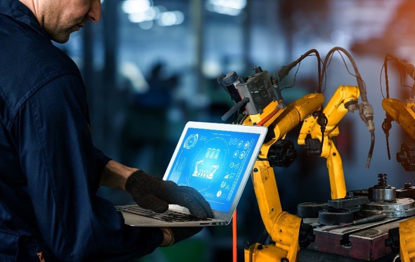 Symbolbild zum Digitalisierungsdarlehen: Mitarbeiter steht am Laptop vor eines Maschinenroboters in einer Produktionshalle